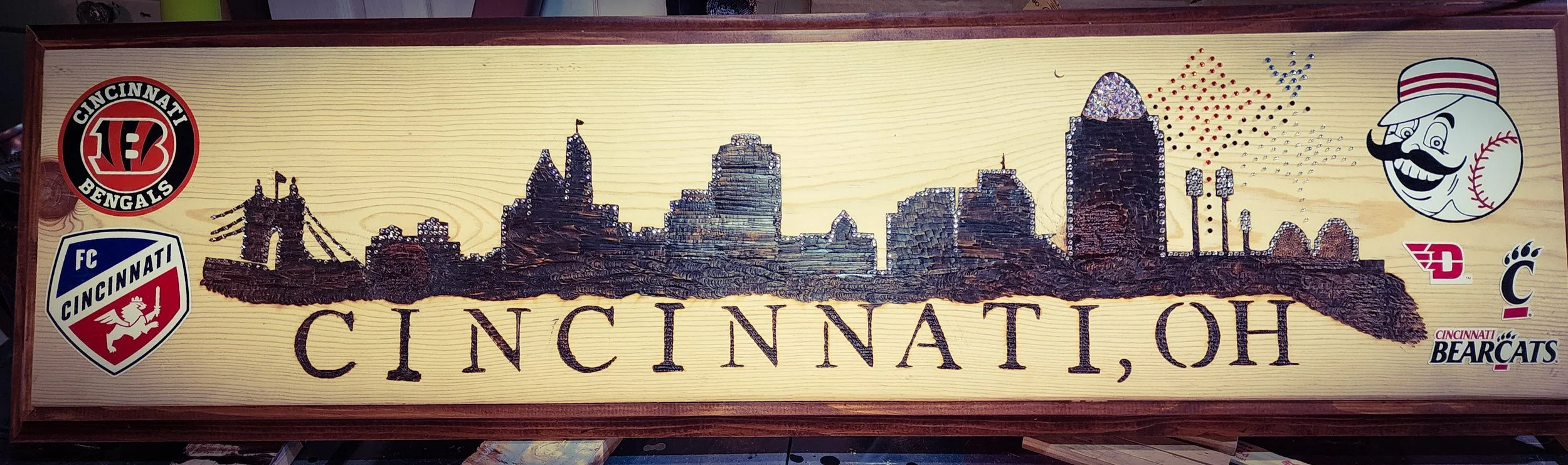 Wooden sign with silhouette of Cincinnati skyline, surrounded by Cincinnati Bengals and Cincinnati Bearcats logos, featuring mascot with baseball and mustache, and cityscape background.