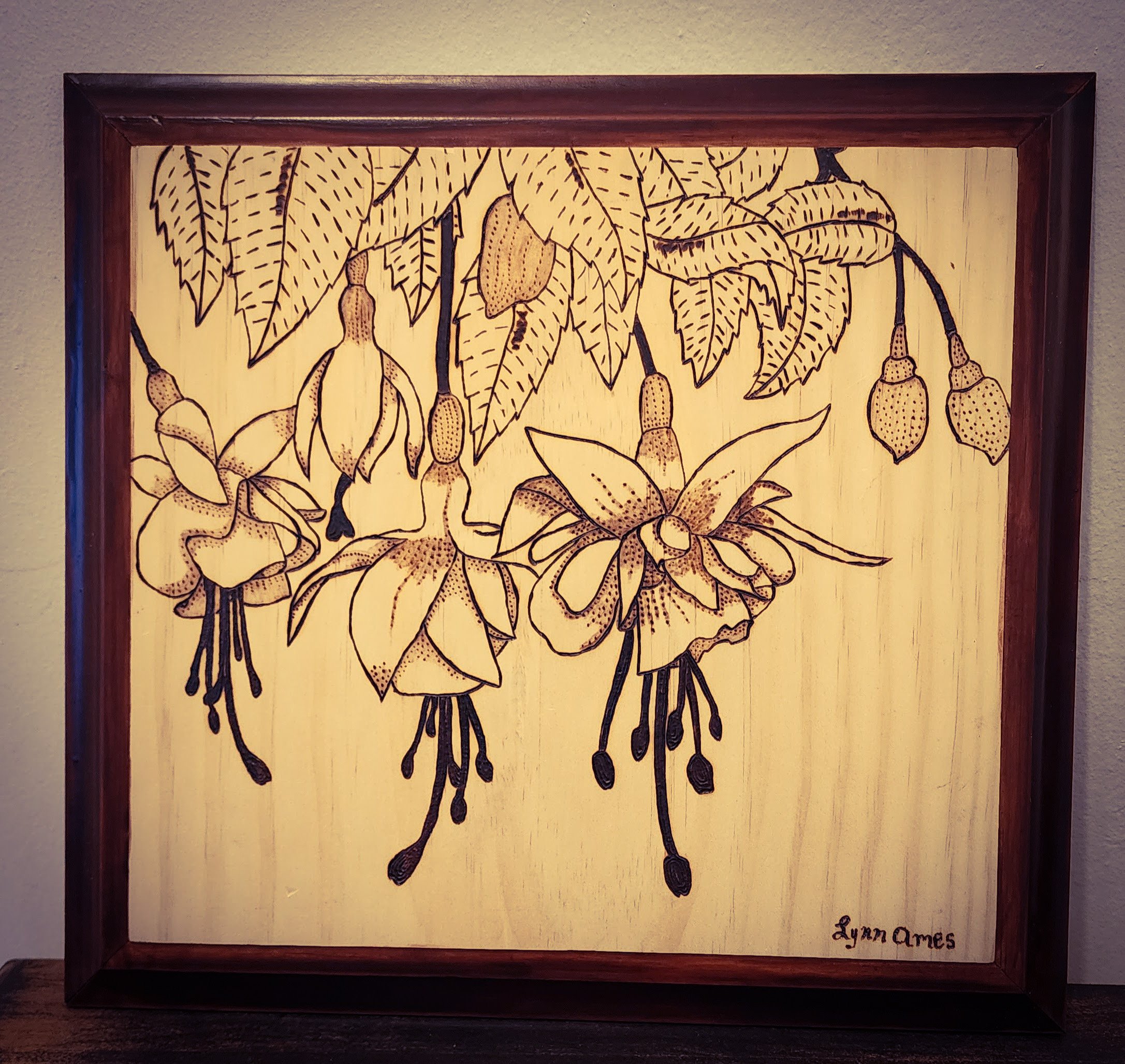 Wood burnt framed artwork featuring a black line drawing of fuchsia flowers and leaves, signed by Lynn Ames.