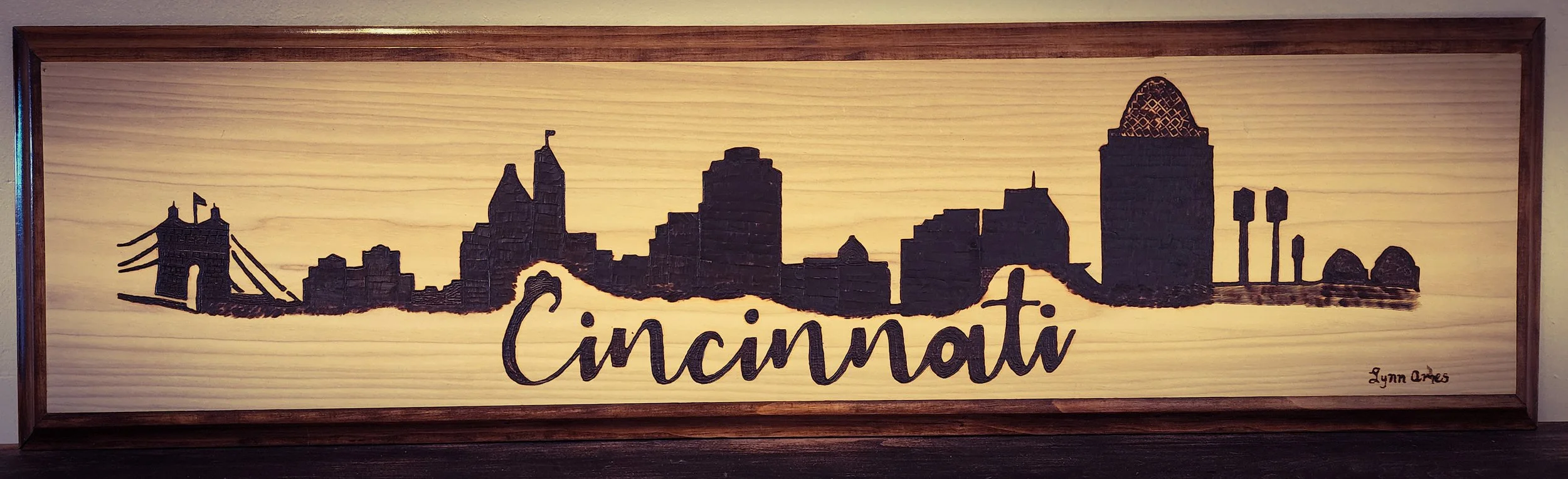 Wooden wall art of the Cincinnati skyline silhouette with the word 'Cincinnati' in cursive script and a small signature 'Lynn Ames' in the bottom right corner.