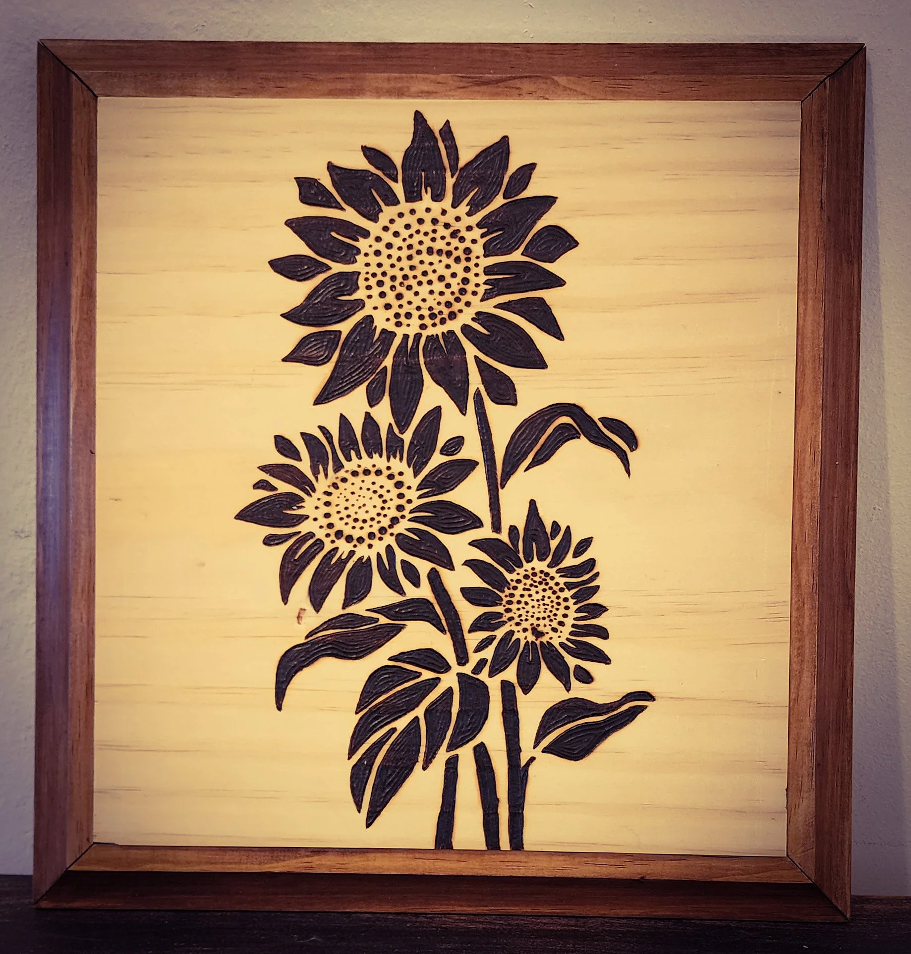 Illuminated wooden art piece depicting black silhouettes of three sunflowers with stems and leaves against a bright background, framed in wood.