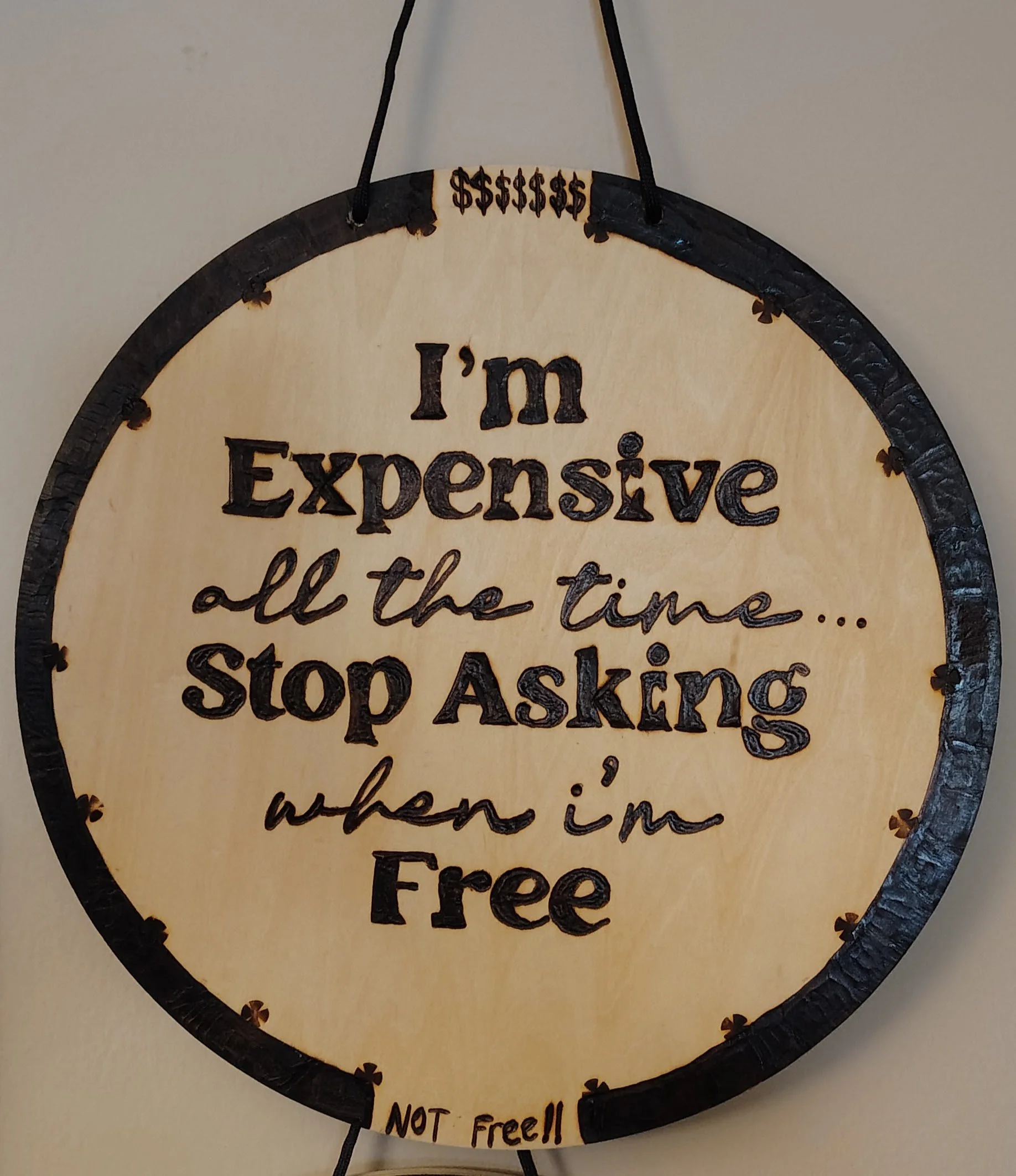 A circular wood burnt sign with black border and hanging cord. The sign has black handwritten-style text that says, 'I'm Expensive all the time... Stop Asking when I'm Free.' At the bottom, in smaller letters, it says, 'NOT free!!'