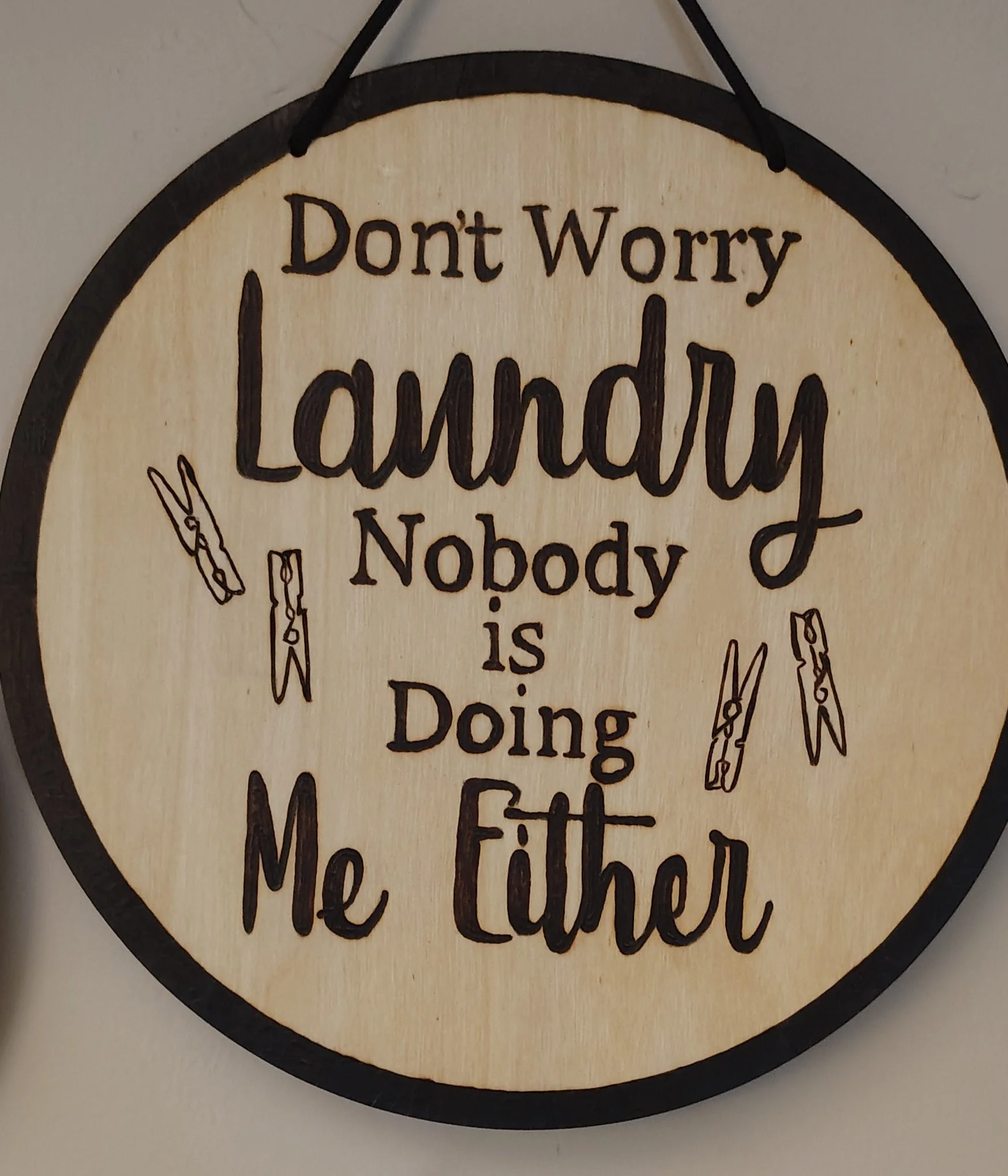 A round wooden sign with black border and black string at the top. It has a humorous message in black text that says, "Don't Worry Laundry Nobody is Doing Me Either," with illustrations of clothespins.