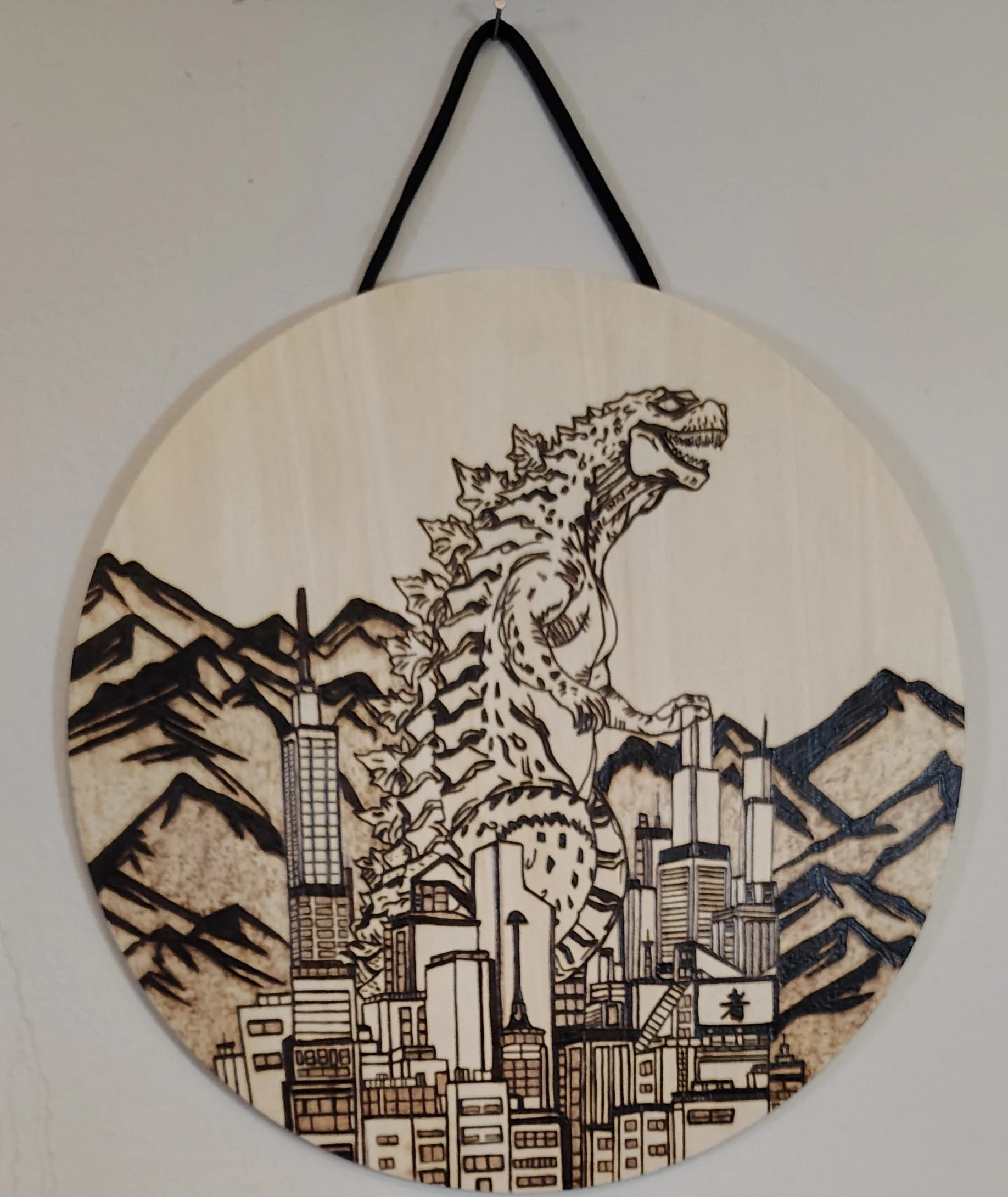 Wood burnt wall hanging depicting a giant monster resembling Godzilla rising over a city with mountains in the background.