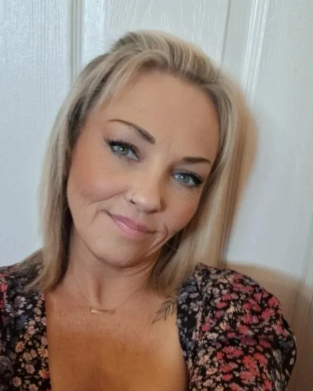 Close-up of a woman with blonde hair, blue eyes, and a gentle smile, wearing a floral top and a delicate necklace, sitting indoors against a white paneled wall.