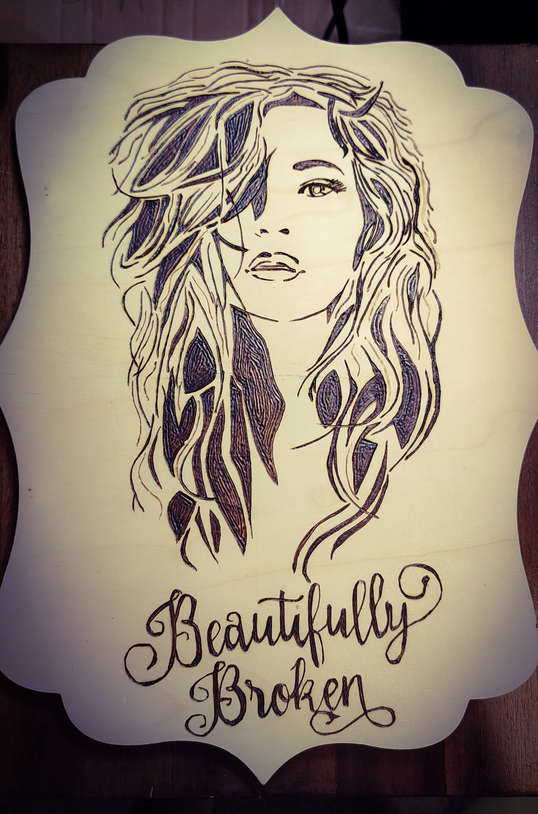 A wooden wall art piece featuring a line drawing of a woman's face with long, wavy hair and the words "Beautifully Broken" written below in cursive.