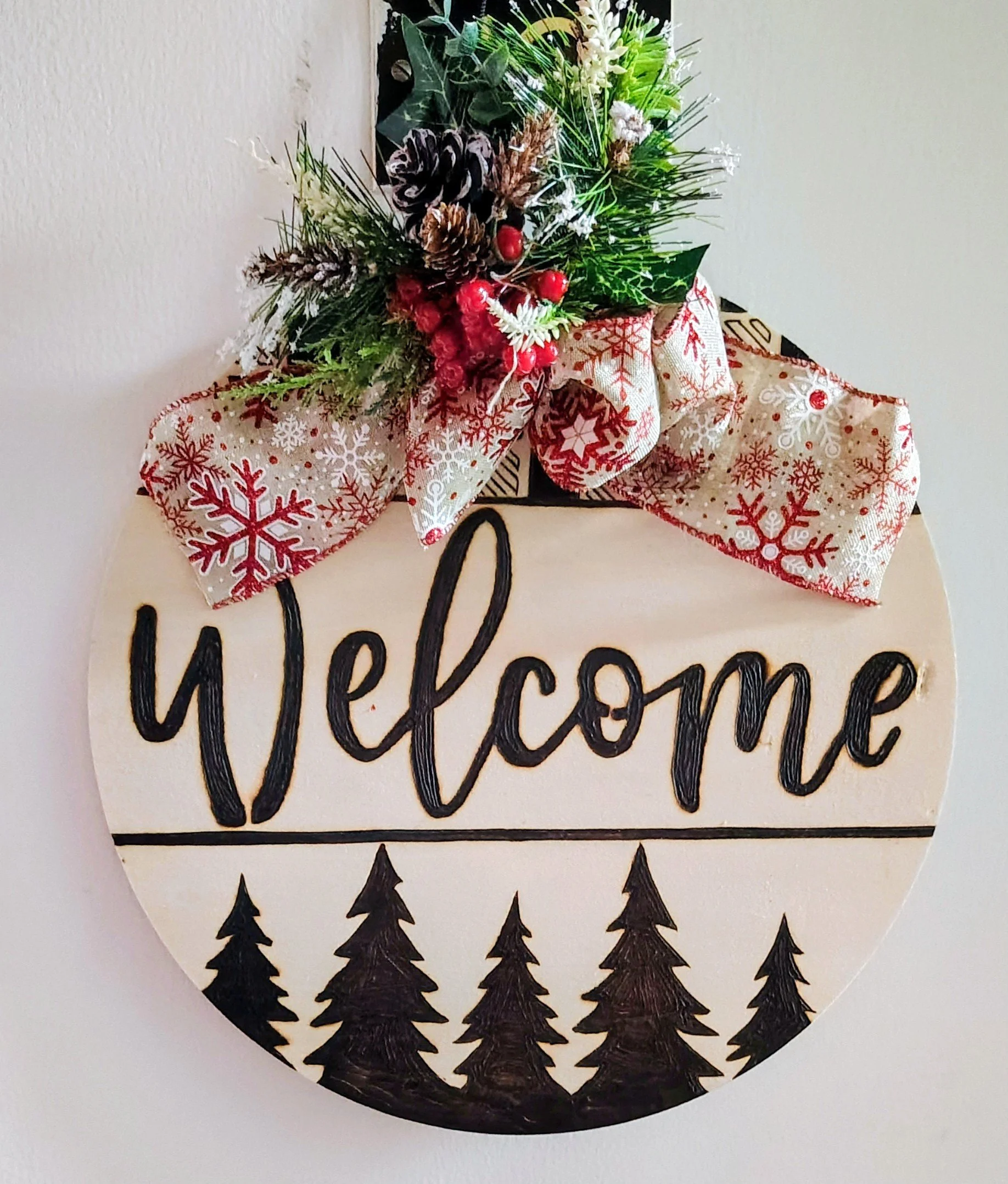 A round wood burnt welcome sign decorated with a Christmas bow and holiday greenery at the top, featuring pinecones, holly with red berries, and a poinsettia, with black wood burnt pine trees at the bottom.