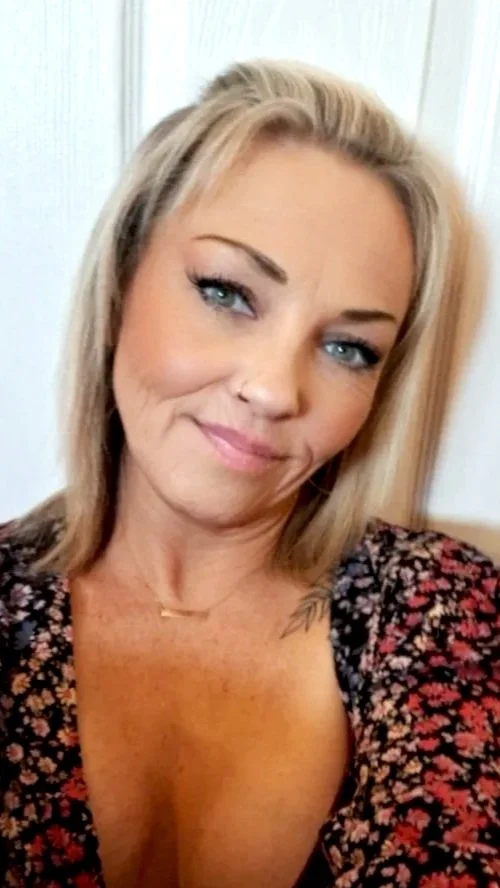 A woman with blonde hair, blue eyes, and a nose piercing, smiling at the camera, wearing a floral top.