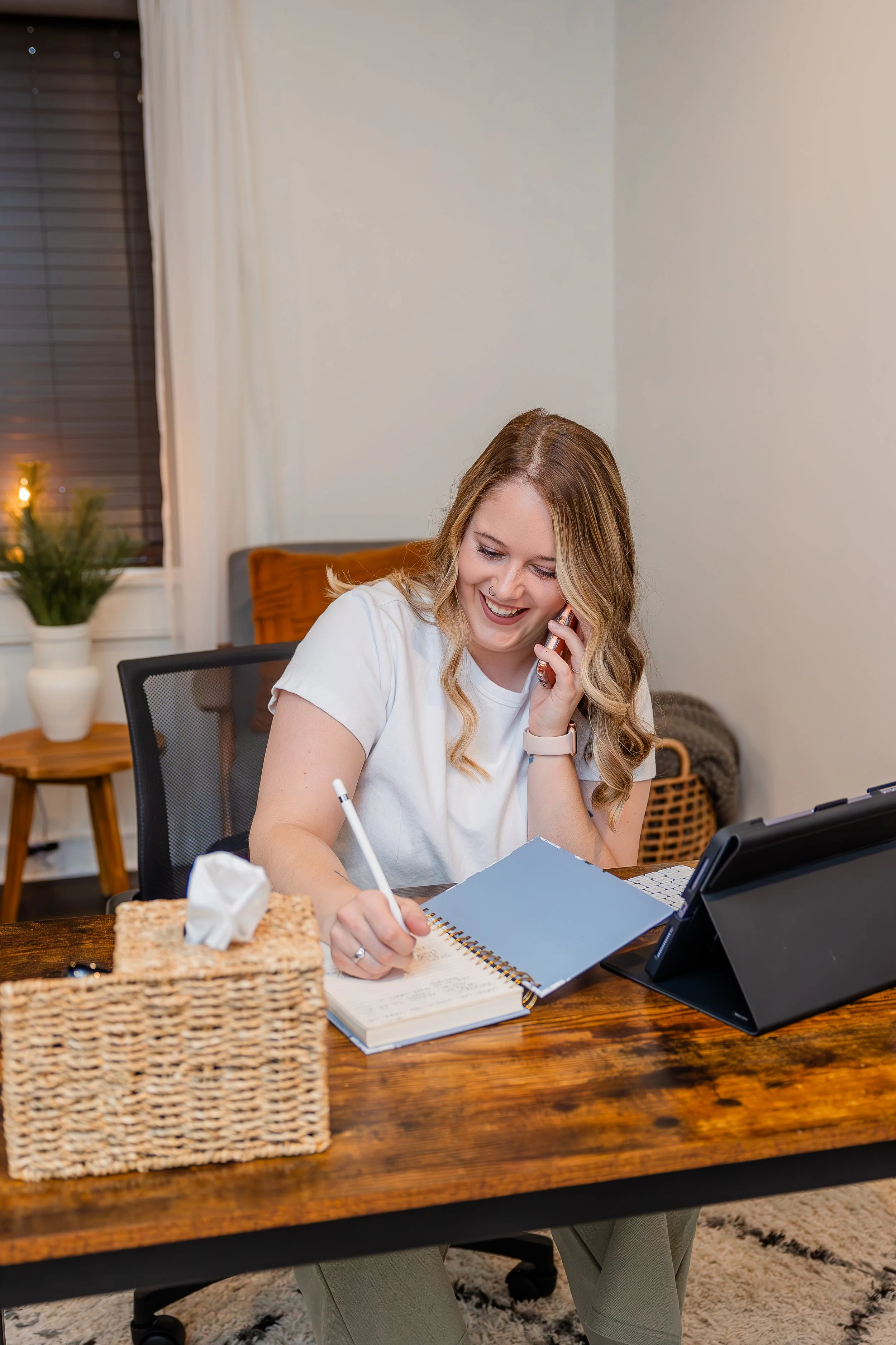 A woman sitting at a wooden desk, smiling while talking on a mobile phone and writing in a notebook, with a tablet device and tissue box on the desk.