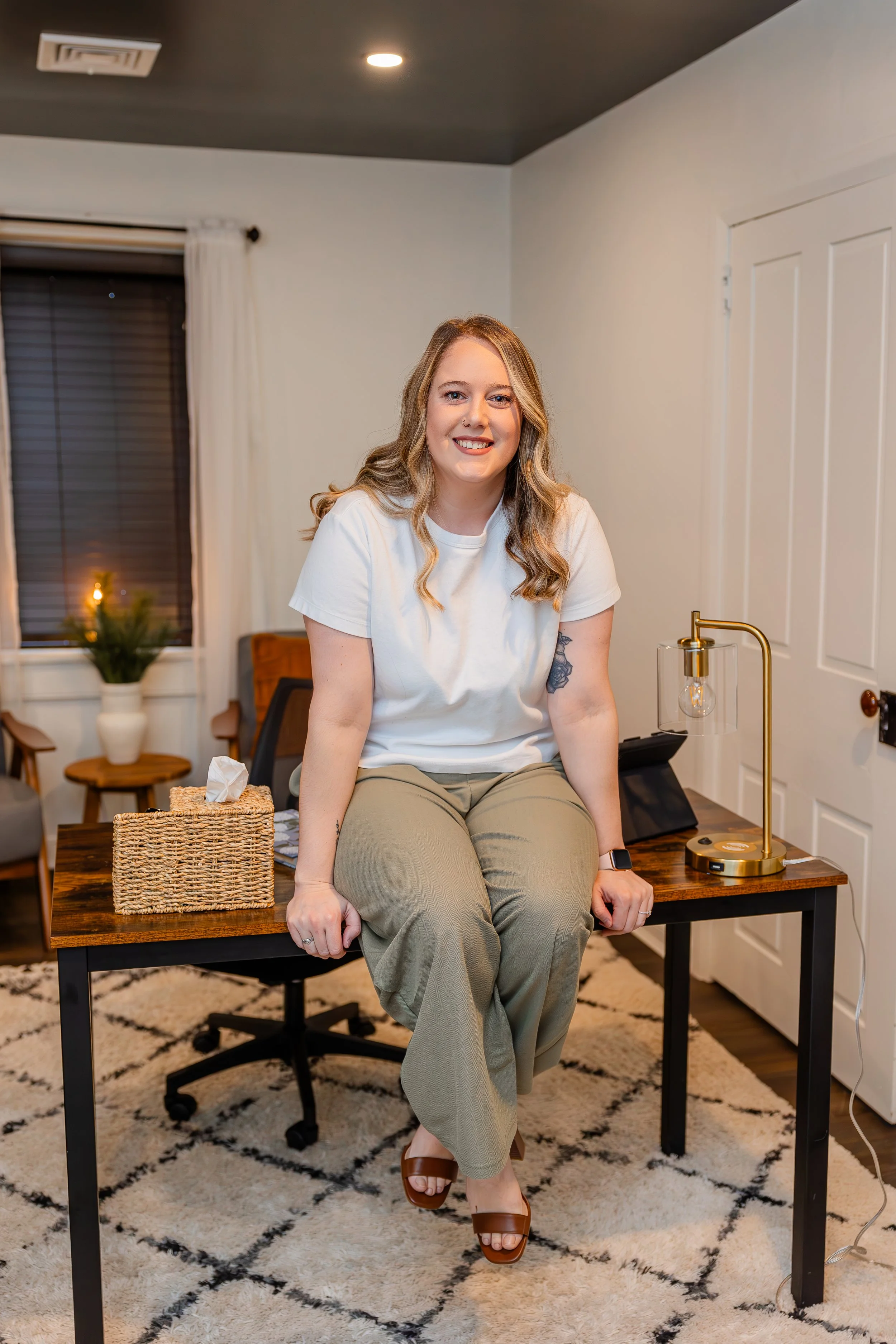 A woman with blonde wavy hair, wearing a white t-shirt and khaki pants, sitting on a wooden desk in a well-lit room with modern decor.
