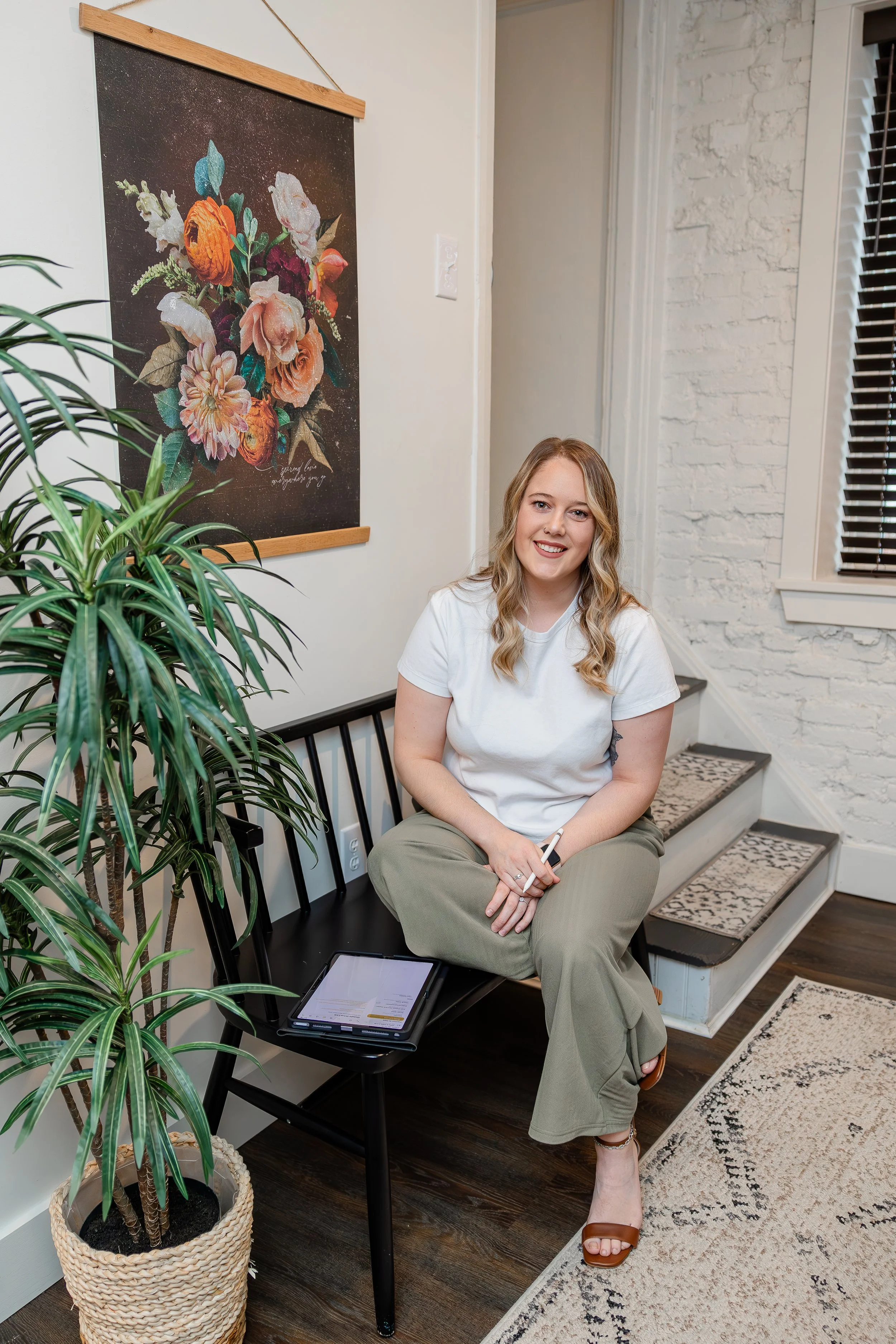 A woman with wavy blonde hair sitting on a black bench in a cozy room with white brick walls, floral artwork hanging on the wall, a potted plant on the left, a window with dark blinds, and a patterned rug on the dark wood floor.