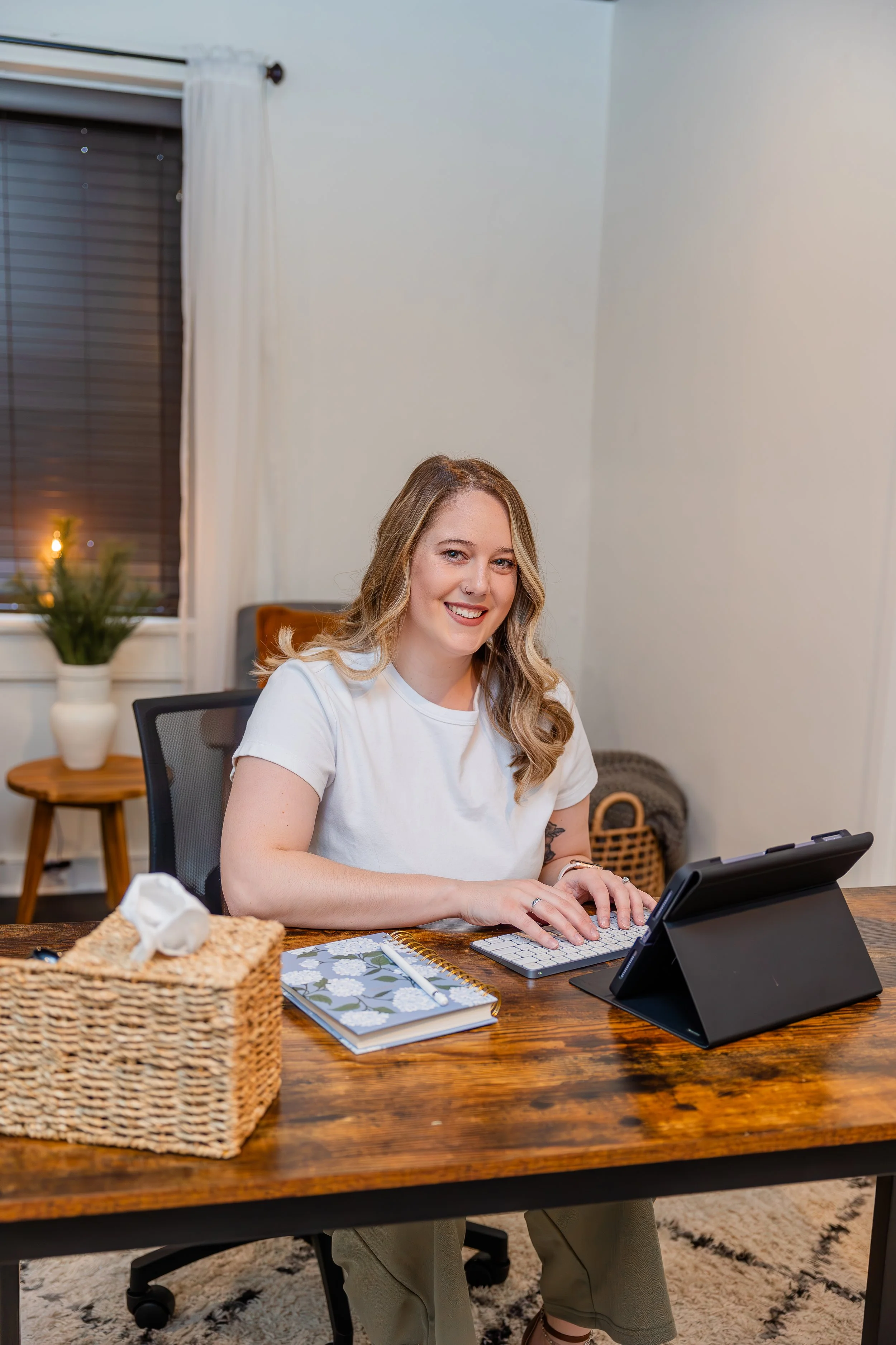 A woman with blonde hair sitting at a wooden desk, smiling and working on a tablet with a keyboard. There is a notebook, a tissue box, and a pen on the desk. A window with white curtains and a potted plant are in the background.