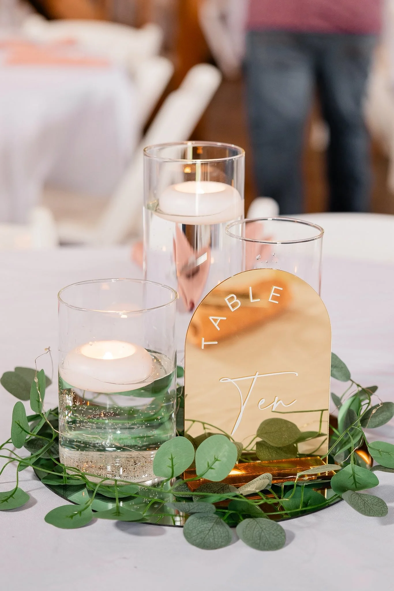 A decorative table centerpiece with three glass candle holders containing floating candles and water, surrounded by green leaves, and a gold sign with the words 'TABLE Ten' on a mirror base.