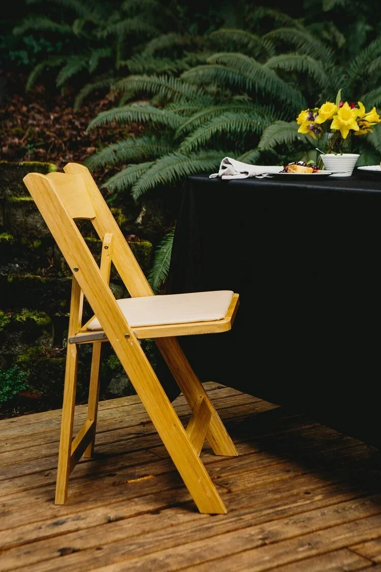 A wooden folding chair with a beige cushion on a wooden deck, next to a black-covered table with a white bowl of yellow flowers and some food, set outdoors with lush green ferns in the background.