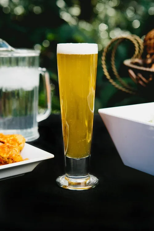 A tall glass of light-colored beer with foam on top, placed on a dark surface outdoors, with a pitcher of water, a plate of food, and a white bowl nearby.