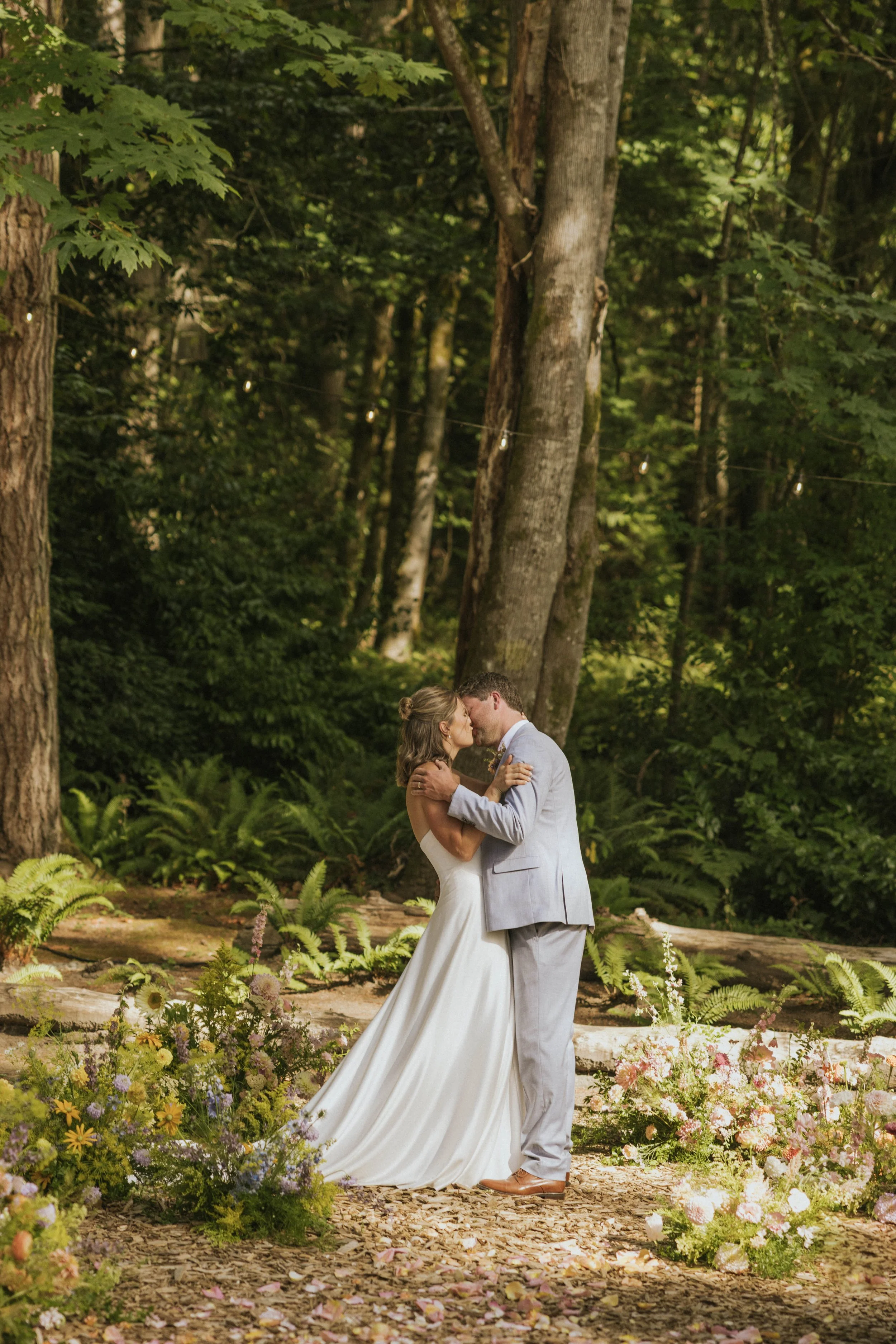 A bride and groom sharing a kiss in a forested outdoor wedding setting surrounded by flowers and greenery.
