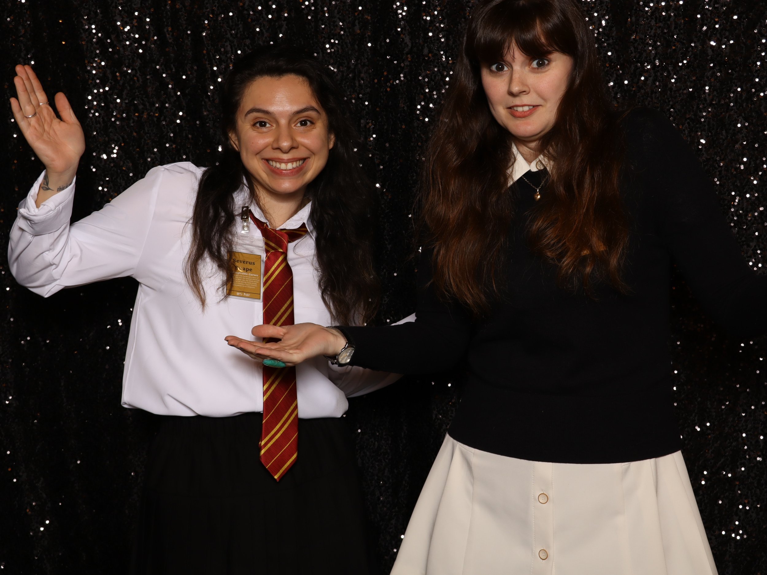 Two young women posing in front of a black sparkly backdrop, one wearing a white shirt and a red striped tie, smiling and raising her right hand, the other wearing a black sweater and white skirt, with an inquisitive expression (Lightning Booth).