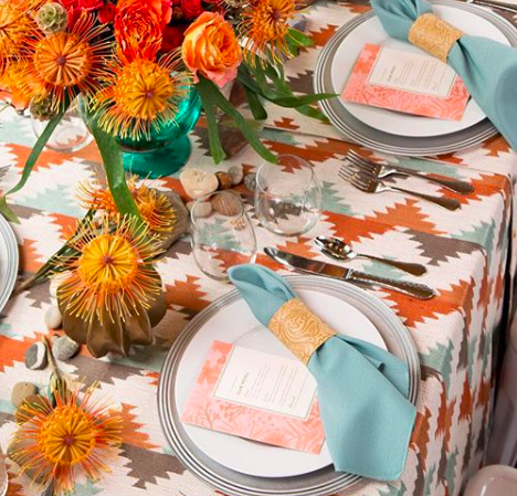 Dining table decorated with a colorful patterned tablecloth, floral centerpiece in a green vase, plates with blue napkins and gold napkin rings, glassware, and cutlery, set for a meal.
