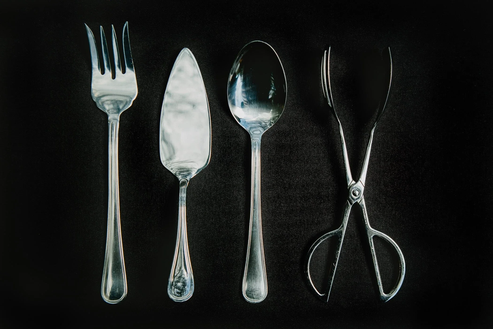 A set of four pieces of cutlery arranged on a black background, including a fork, two knives, a spoon, and a pair of scissors.