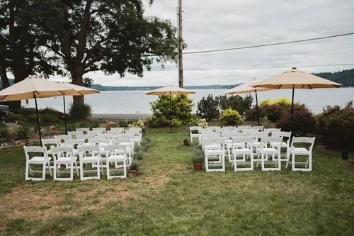 Outdoor seating arrangement with white chairs and beige umbrellas on a grassy area by a body of water, prepared for an event.