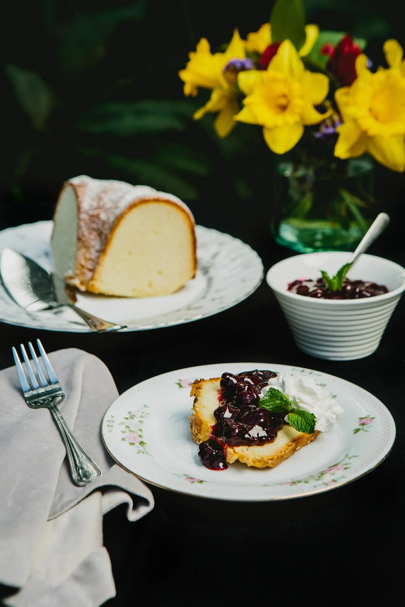 A slice of cheesecake topped with berry compote, served with whipped cream and mint on a floral plate, with a fork and napkin nearby. In the background, a whole cheesecake and a bowl of berry jam, with yellow flowers in a vase.