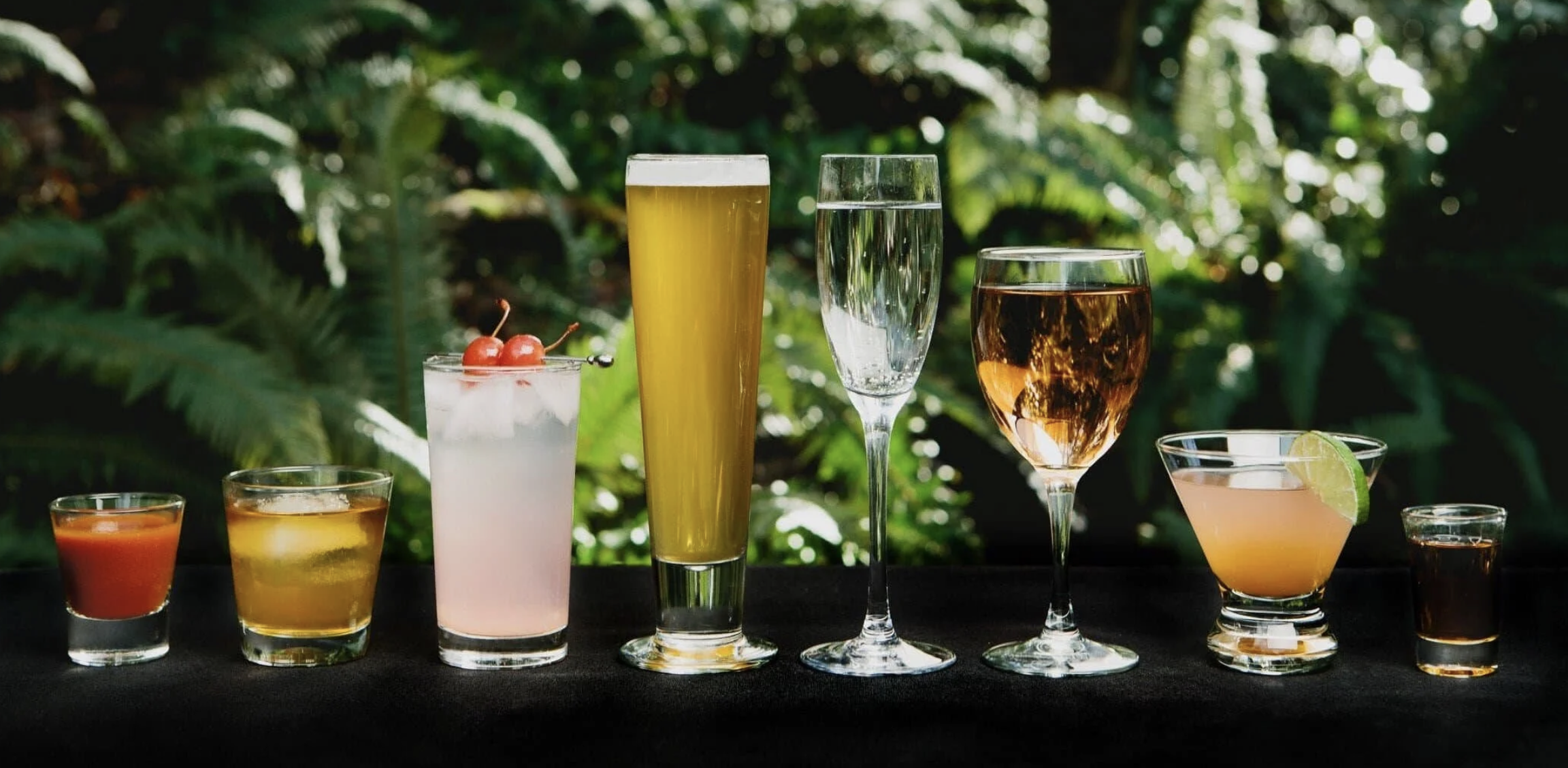 A variety of colorful cocktails and drinks arranged on a black table with a lush green outdoor background.