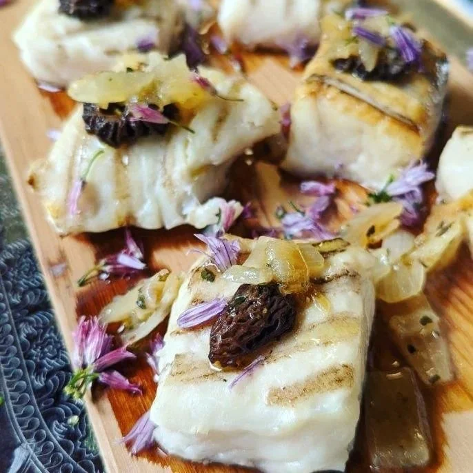 Cooked fish fillets topped with chopped onions, black garlic, and purple flower petals on a wooden platter.
