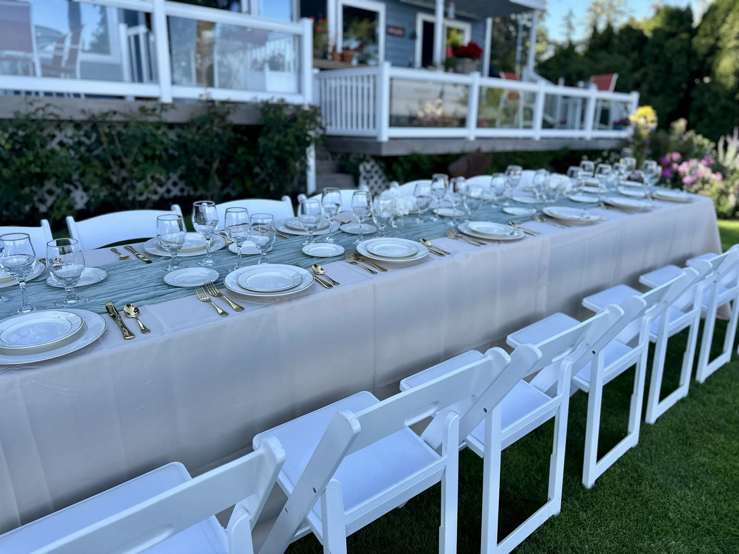Long outdoor table set for a gathering with white plates, silverware, and glasses, surrounded by white chairs, in a garden with a house featuring a porch and flowers in the background.