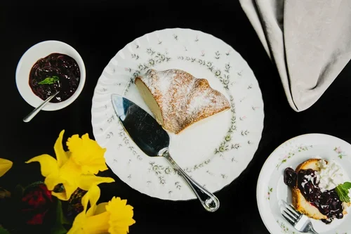A slice of cake with powdered sugar on a decorative white plate, a small bowl of dark berry jam with a spoon, a cupcake with dark berry topping on a small white plate, yellow flowers, and a white napkin.