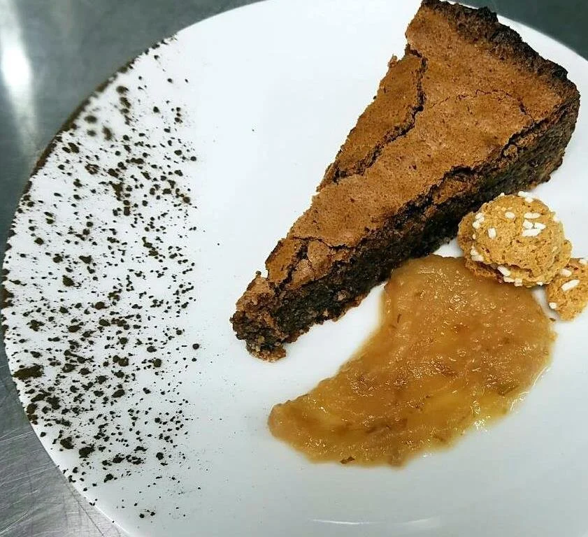Slice of chocolate brownie on a white plate, garnished with a dusting of cocoa powder on one side, with apple sauce and a small cookie on the side.