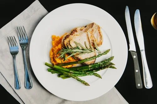 Plate with grilled chicken breast, mashed sweet potatoes, and grilled asparagus, garnished with rosemary, on a table with cutlery and a napkin.