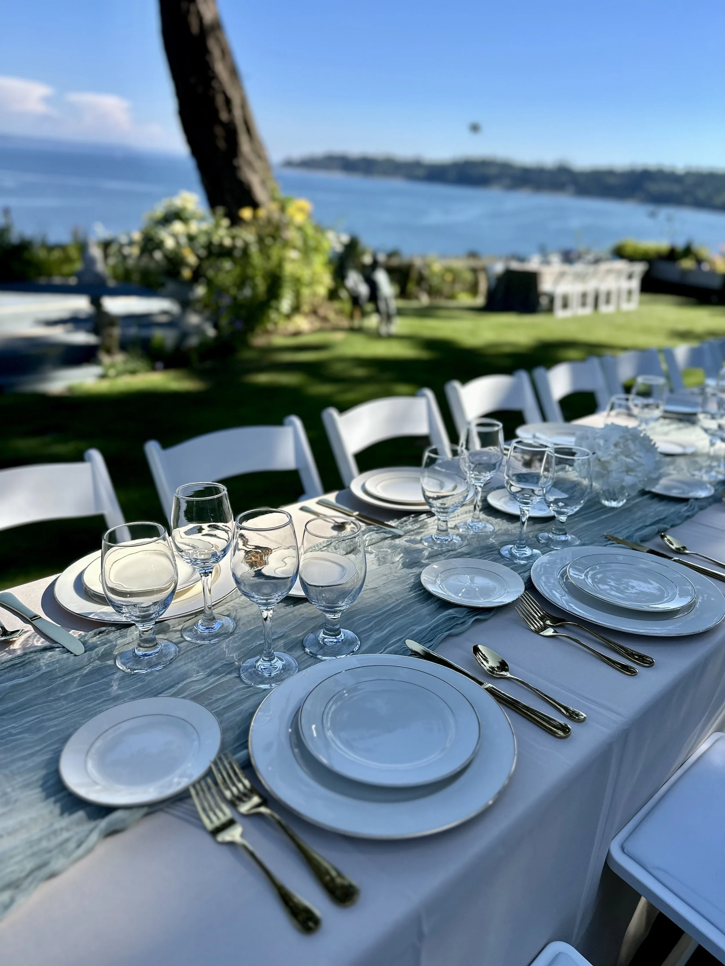 Elegant outdoor dining table set with white plates, silverware, water glasses, and a floral centerpiece, overlooking a scenic view of the ocean and land.