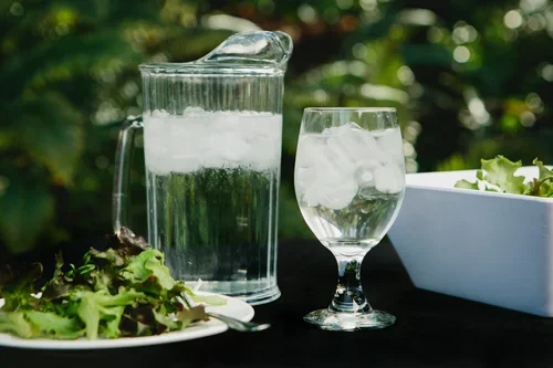 A glass pitcher of water with ice, a glass of ice water, a salad, and a bowl of greens on a dark outdoor table.