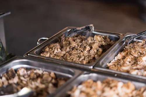 Multiple stainless steel food trays filled with cooked ground meat, with tongs resting on top of one tray.