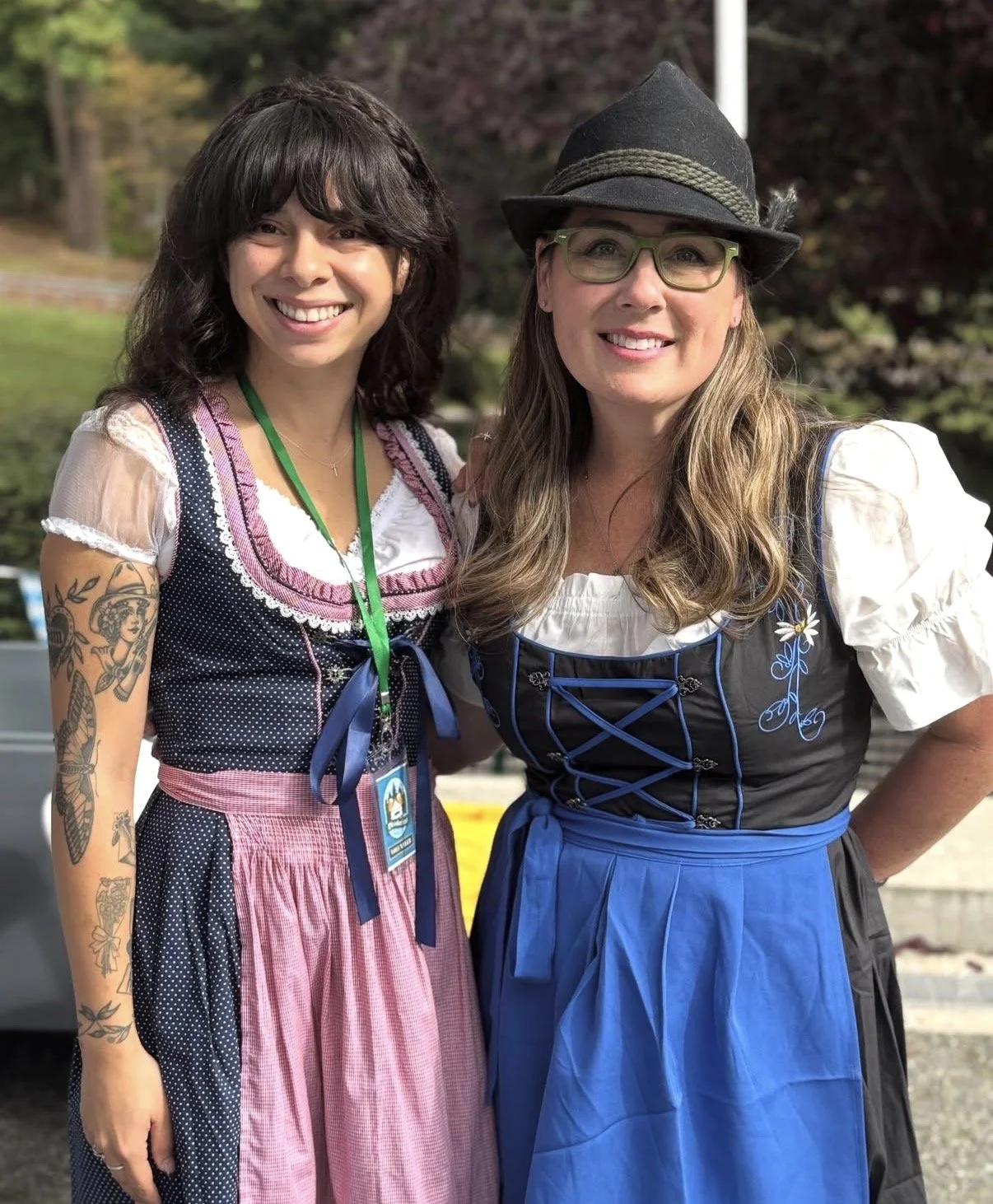 Two women wearing traditional German dirndl dresses standing outdoors, smiling at the camera.
