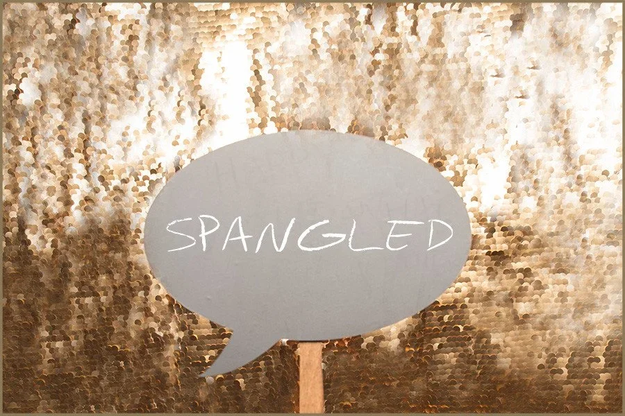 A speech bubble made of paper with the word 'SPANGLED' written on it, standing in front of a gold, sequined backdrop.