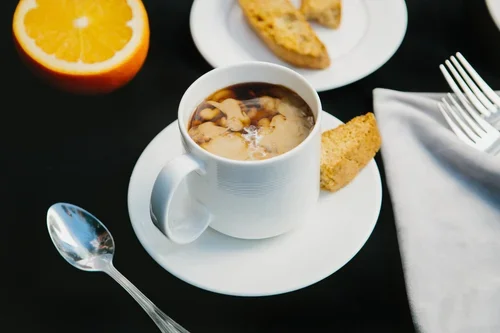 Cup of mushroom soup on a white saucer with a slice of bread, an orange half, and a fork and spoon on a black table.