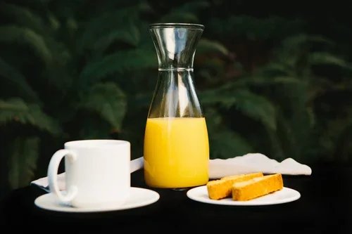 A glass pitcher filled with orange juice, a white coffee cup on a saucer, and a small plate with two waffles, all set on a black surface with a dark green leafy background.