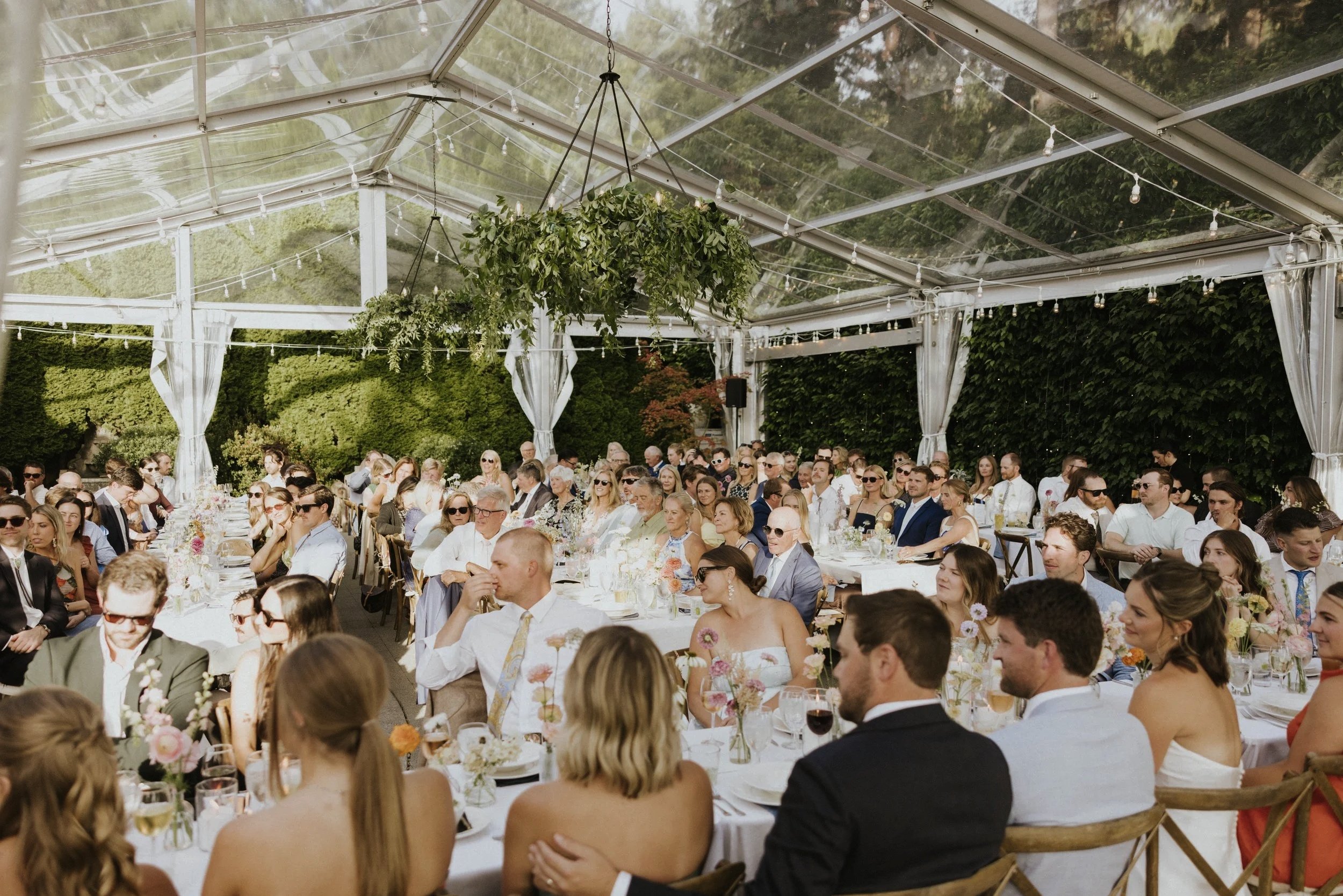 A large outdoor wedding reception under a transparent tent with string lights and greenery. Guests seated at decorated tables, some wearing sunglasses, enjoying food and drinks.