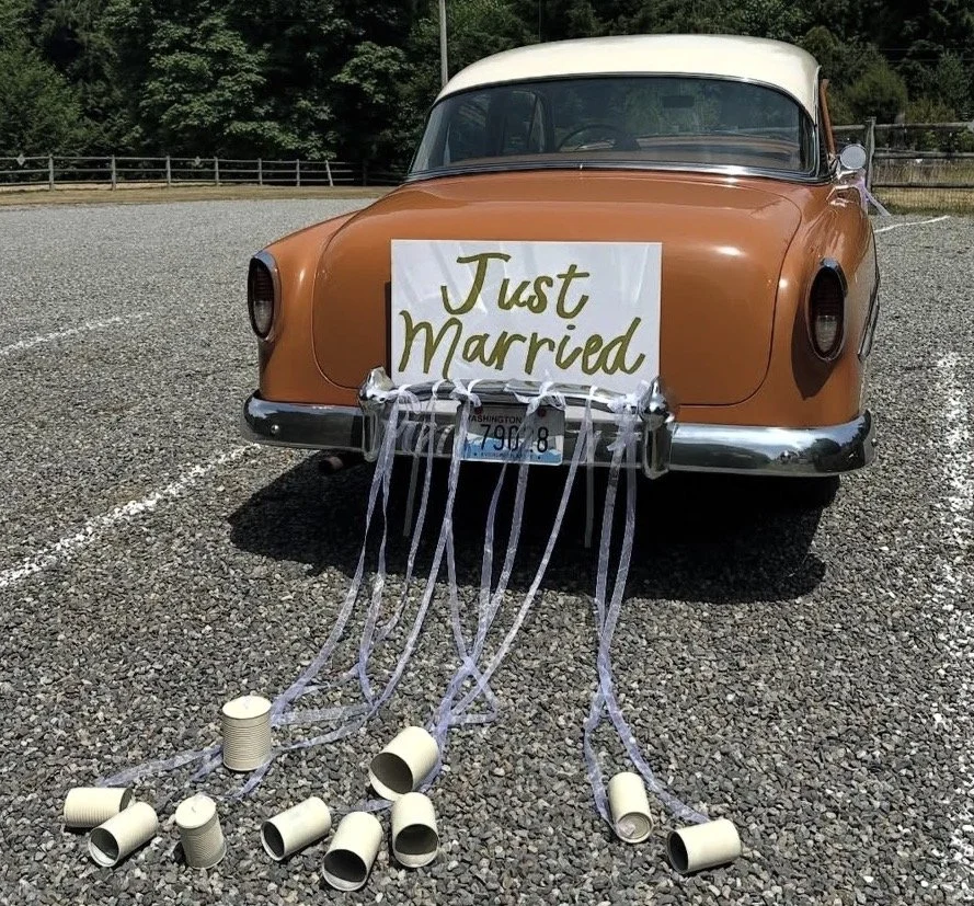 A vintage orange and white car with a sign on the front that reads 'Just Married.' White ribbons and empty paper cups are attached to the bumper, with the cups spilled onto the gravel ground.