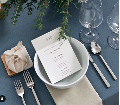 Elegant table setting with a menu inside a white bowl, two wine glasses, silverware, a beige napkin, a small bouquet of greenery, and a wrapped gift on a blue tablecloth.