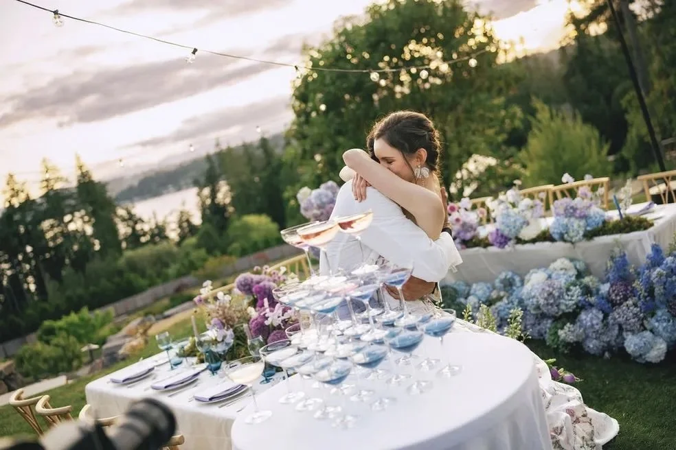 A woman in a wedding dress hugs a man at an outdoor wedding reception with a decorated table and a scenic background of trees and a lake at sunset.