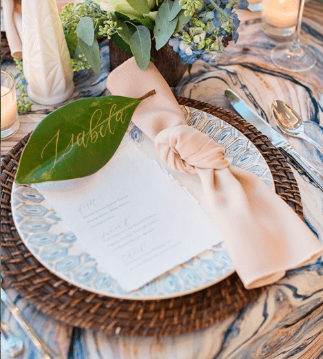 Elegant table setting with a decorative blue and white patterned plate on a woven charger, a blush napkin, a large green leaf with gold writing, a menu card, a floral arrangement, a glass of beverage, and cutlery.