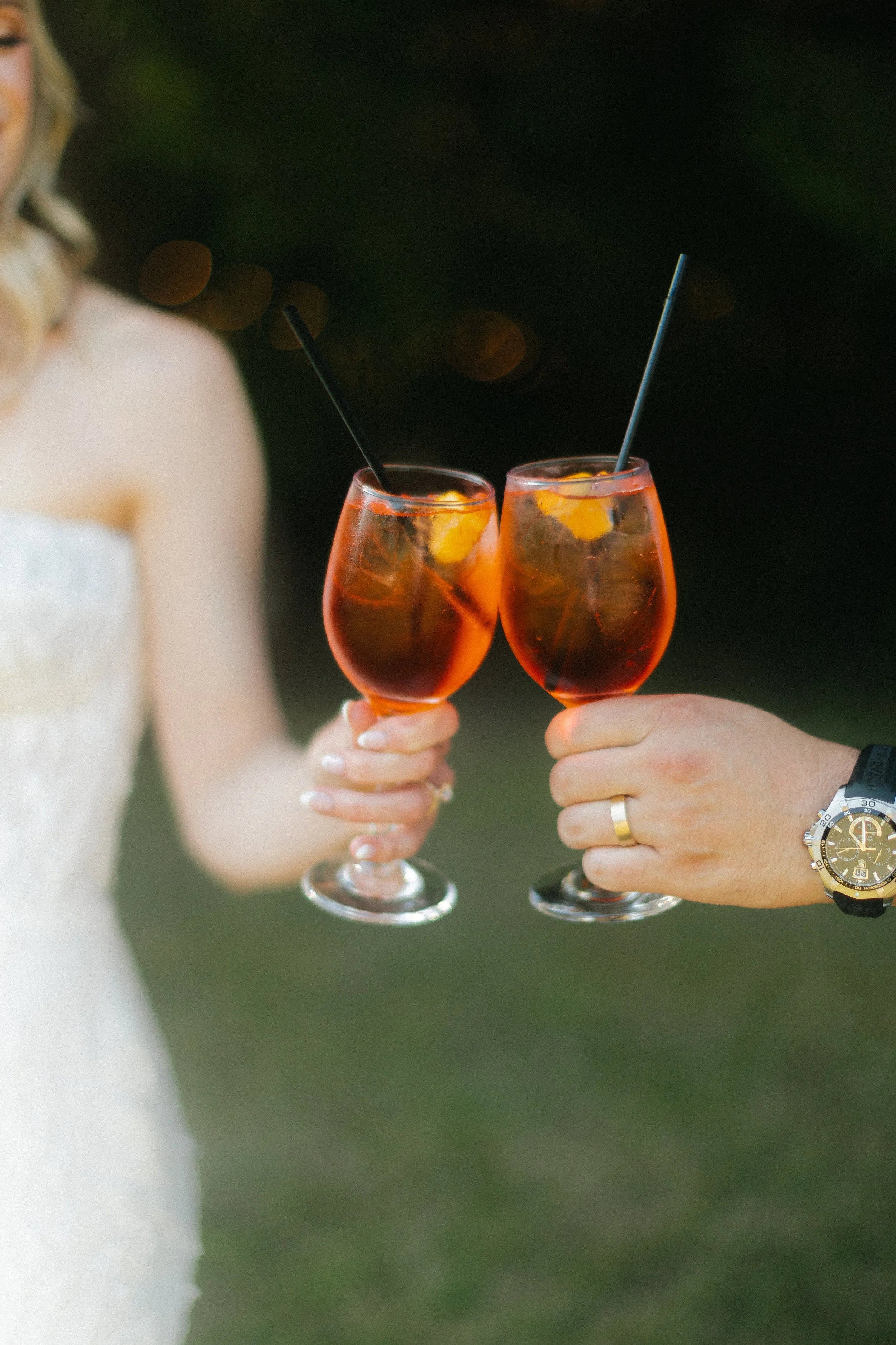 Two people clinking glasses of dark red cocktails with lemon slices, outdoors at night.