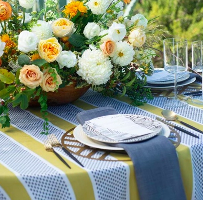 A table set for an outdoor meal with a floral centerpiece of white, cream, and peach flowers, and tableware including plates, glassware, and cutlery, all arranged on a yellow and white striped tablecloth.