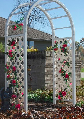 White garden arch with hanging pink roses, situated in a flower garden with trees and a house in the background.