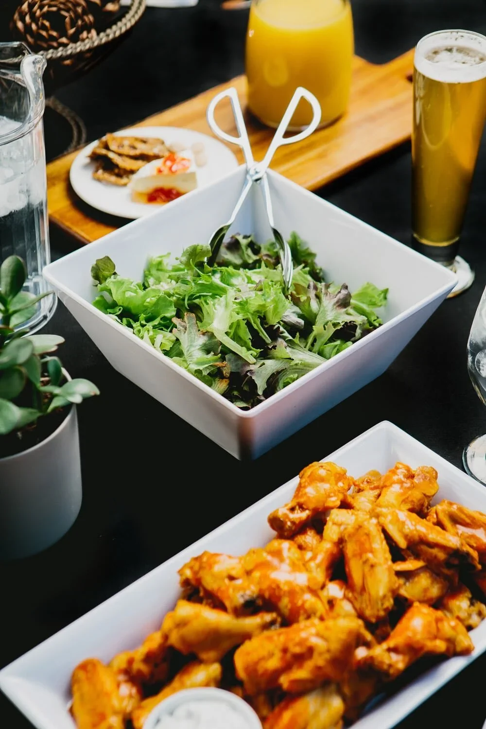 A table with a bowl of mixed green salad, fried chicken wings, and drinks including beer and orange juice.