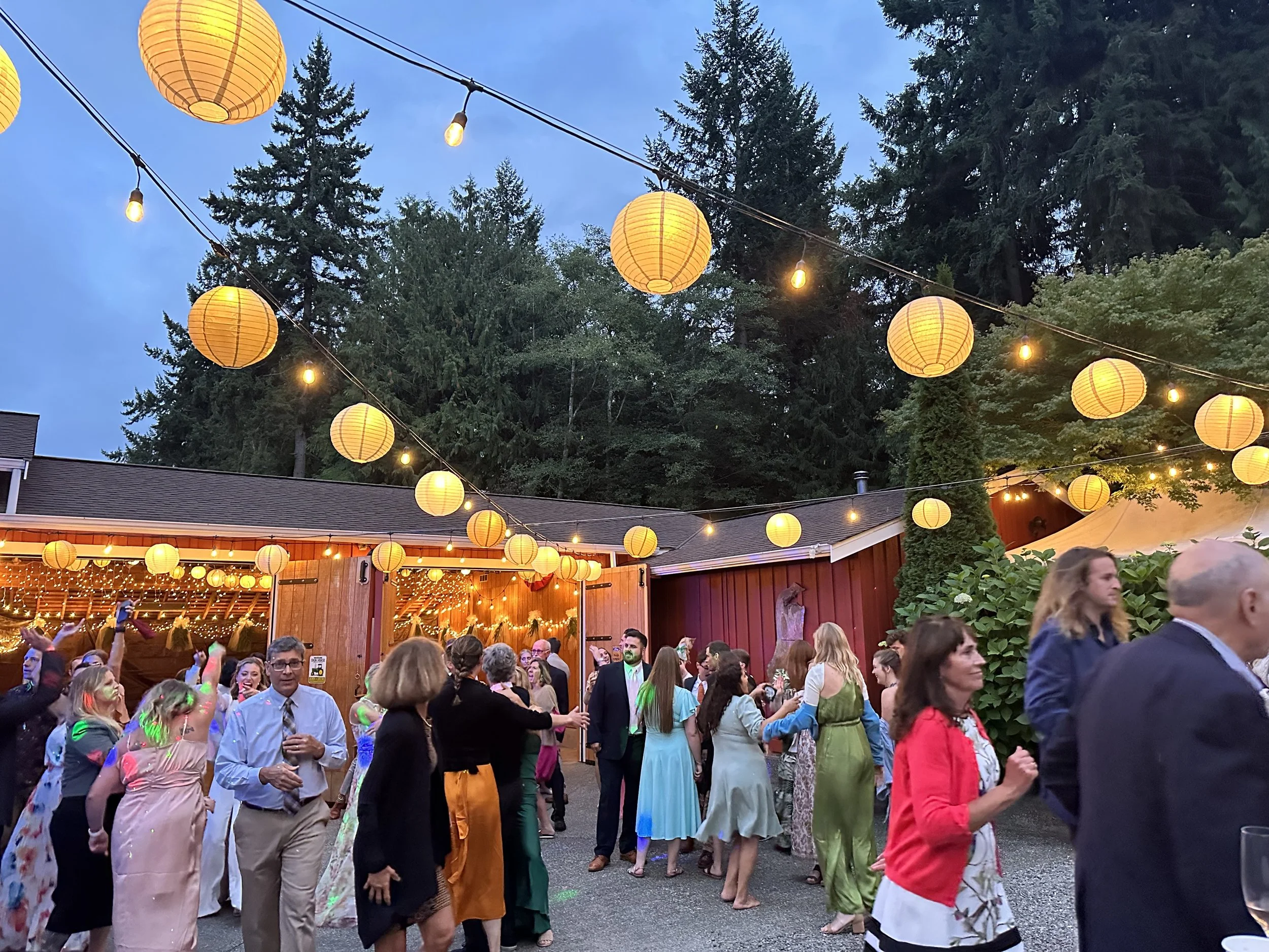 People dancing and socializing outdoors at evening under hanging paper lanterns and string lights, with trees in the background.