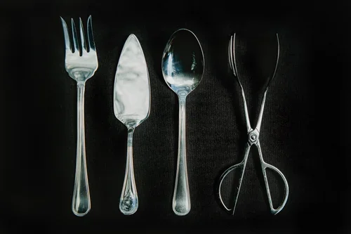 A set of four metal kitchen utensils on a black surface: a fork, a spatula, a spoon, and a pair of tongs.