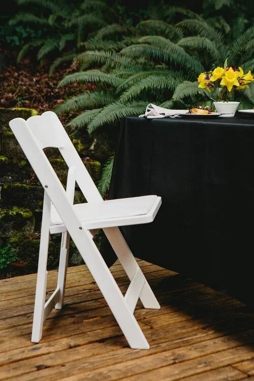 A white folding chair next to a black table with a flower arrangement of yellow and purple flowers, set outdoors on a wooden deck with green ferns in the background.