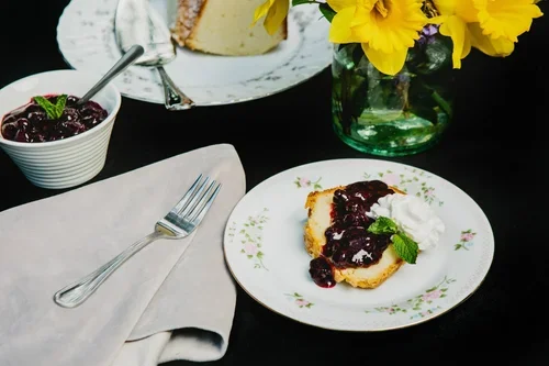 A plate with a slice of cheesecake topped with cherry compote and a mint leaf, a bowl of cherry compote with a spoon, a fork on a napkin, a vase of yellow flowers, and a whole cheesecake in the background.